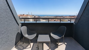 A balcony with two chairs and a small table, overlooking a coastal cityscape with buildings and a clear sky.