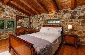 Un dormitorio con pared de piedra, vigas de madera en el techo, una cama de madera, una mesita de noche, un aparador y una ventana con contraventanas.