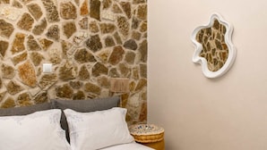 A bedroom with a stone wall, a bed with white linens, a gray pillow, a small round table with a decorative item, and a wall-mounted mirror.