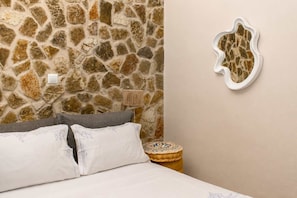 A bedroom with a stone wall, a bed with white linens, a gray pillow, a small round table with a decorative item, and a wall-mounted mirror.