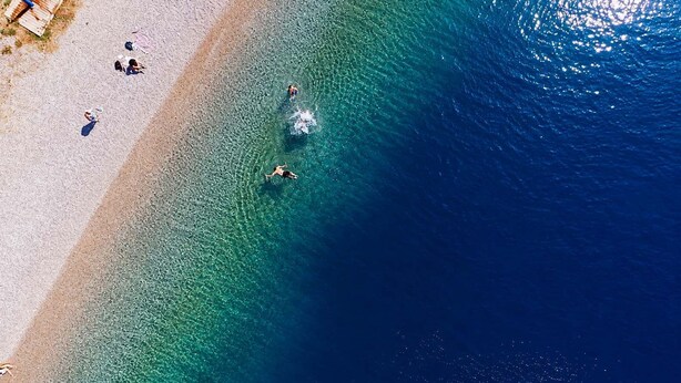 An aerial view of a beach with clear water, a sandy shore, and a few people swimming and sunbathing.