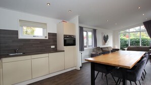 A modern kitchen with a built-in oven, a dining area with a wooden table and chairs, and large windows offering a view of greenery outside. - Holiday Home in Brunssum With Terrace (Brunssum)