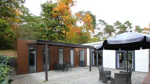 A modern outdoor patio with a brick floor, a covered seating area, and a wooden building with glass doors. - Holiday Home in Brunssum With Terrace (Brunssum)