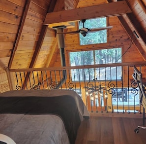 A wooden cabin with a loft bedroom, a ceiling fan, and a view of the forest.
