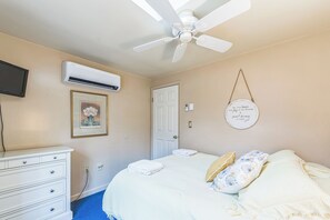 Ein Schlafzimmer mit Bett, Deckenventilator, Klimaanlage, Fernseher, einem gerahmten Bild, einer Tür und einem an der Wand befestigten Schild.