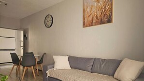 A living room with a grey sofa, a small round table, and two chairs. A clock and a painting of wheat stalks are on the wall. - 1st Generation style (City of Johannesburg Metropolitan Municipality)