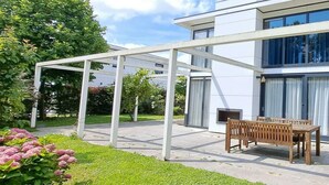 Ein modernes Haus mit überdachter Terrasse, einer Holzbank und einem Rasen.