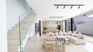 A modern living room with a sofa, coffee table, and a staircase with glass railings. - New 4 Bedroom Villa With Pool, 100 Meters From the Beach (Paralimni)