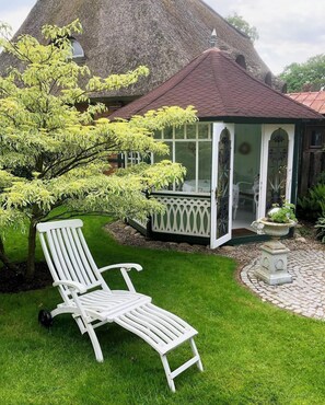 Une maison à toit de chaume, dotée d’un porche abrité, d’une chaise blanche et d’un arbre aux feuilles jaunes.