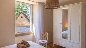 A bedroom with two beds, a wardrobe, a chair, and a window with a view of a house.