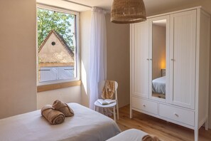 A bedroom with two beds, a wardrobe, a chair, and a window with a view of a house.