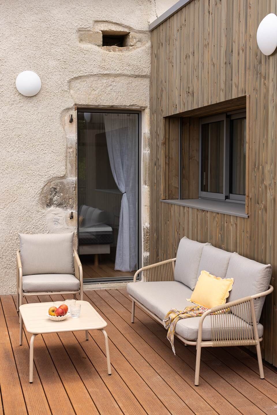 A wooden deck with a wicker sofa and a chair, a small table with fruit, and a window with curtains.