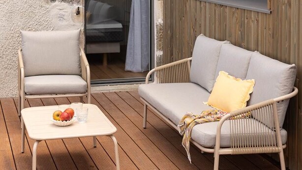 A wooden deck with a wicker sofa and a chair, a small table with fruit, and a window with curtains.