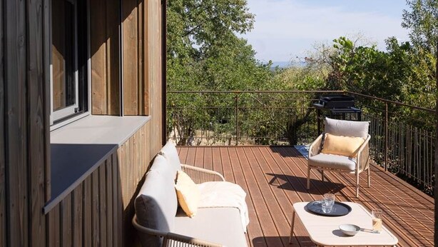 Una terraza al aire libre con piso de madera, dos sillas blancas acolchadas, una mesita y vistas a la vegetación circundante.