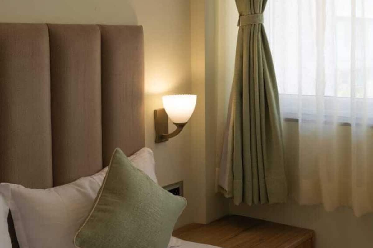 A hotel room with a bed, pillows, a nightstand, a lamp, and a window with curtains.