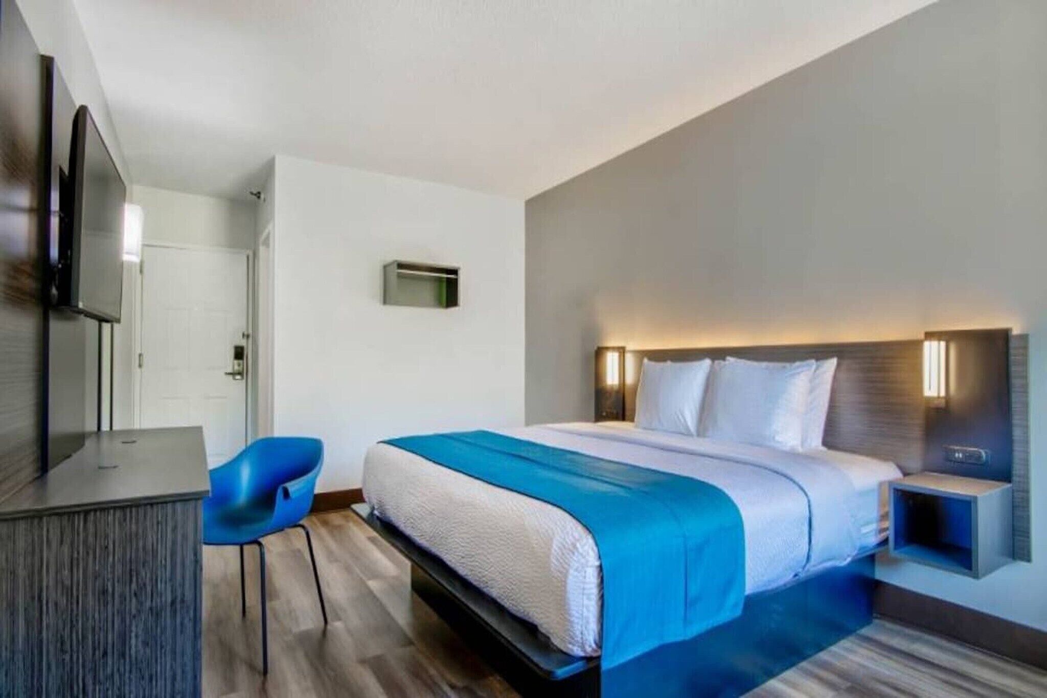 A hotel room with a bed, a desk, a chair, a TV, and a wall-mounted light.
