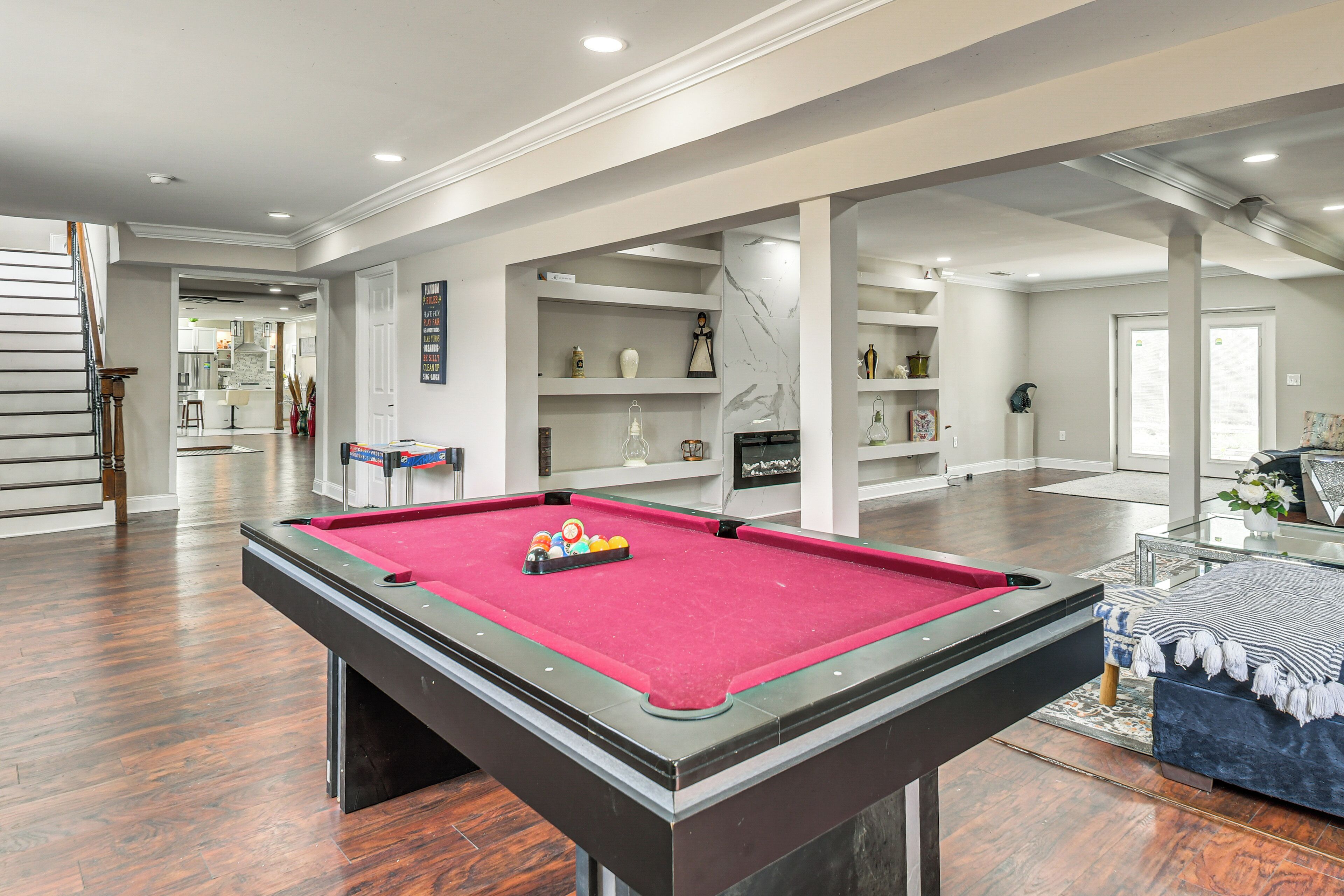 A pool table in a spacious room with a wooden floor, a staircase, and a view into another room.