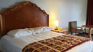 A hotel room with a wooden headboard, a bed with white linens, a patterned throw blanket, a bedside lamp, a chair, and a small table with a telephone.