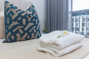 Een net opgemaakt bed met een gekleurd kussen, een stapel witte handdoeken en een klein bloemetje.