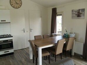 A dining area with a wooden table and chairs, a kitchen with a stove and oven, a clock on the wall, and a window with curtains. - Holiday Home in Dordrecht With Terrace (Dordrecht)