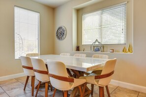 Uma sala de jantar com uma mesa retangular rodeada por seis cadeiras, janelas amplas com persianas e um relógio de parede decorativo.
