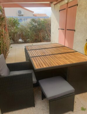 Une terrasse extérieure avec une table en bois et des meubles en osier, entourée de plantes et d’un bâtiment doté d’un toit rouge.