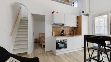 Un ambiente living compatto con angolo cottura, zona pranzo e una scala che conduce a un piano superiore.
