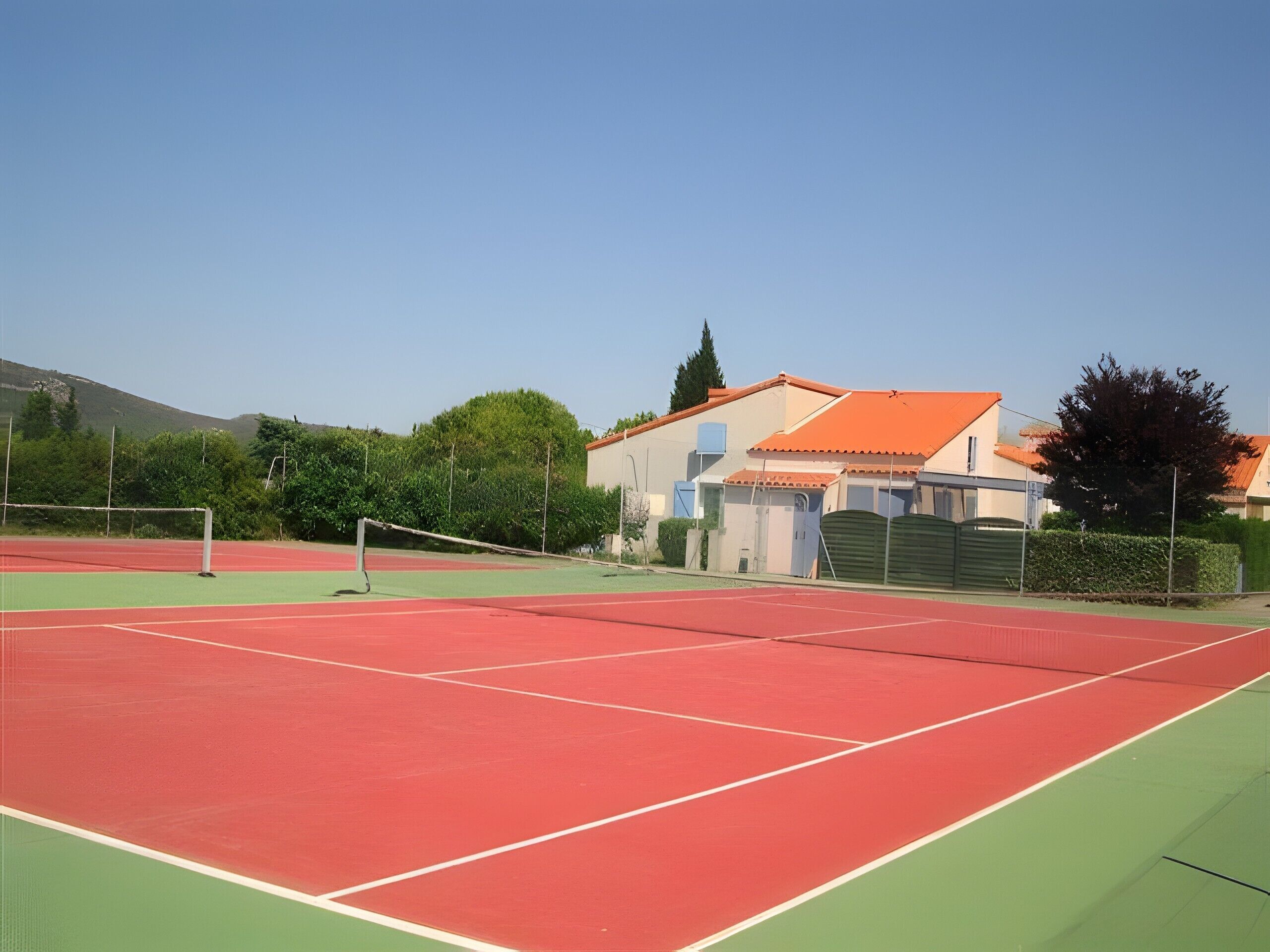 Una cancha de tenis con red, rodeada de un área verde, y un edificio con techo rojo al fondo.