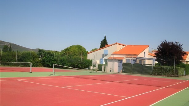 A tennis court with a net, surrounded by a green area, and a building with a red roof in the background.