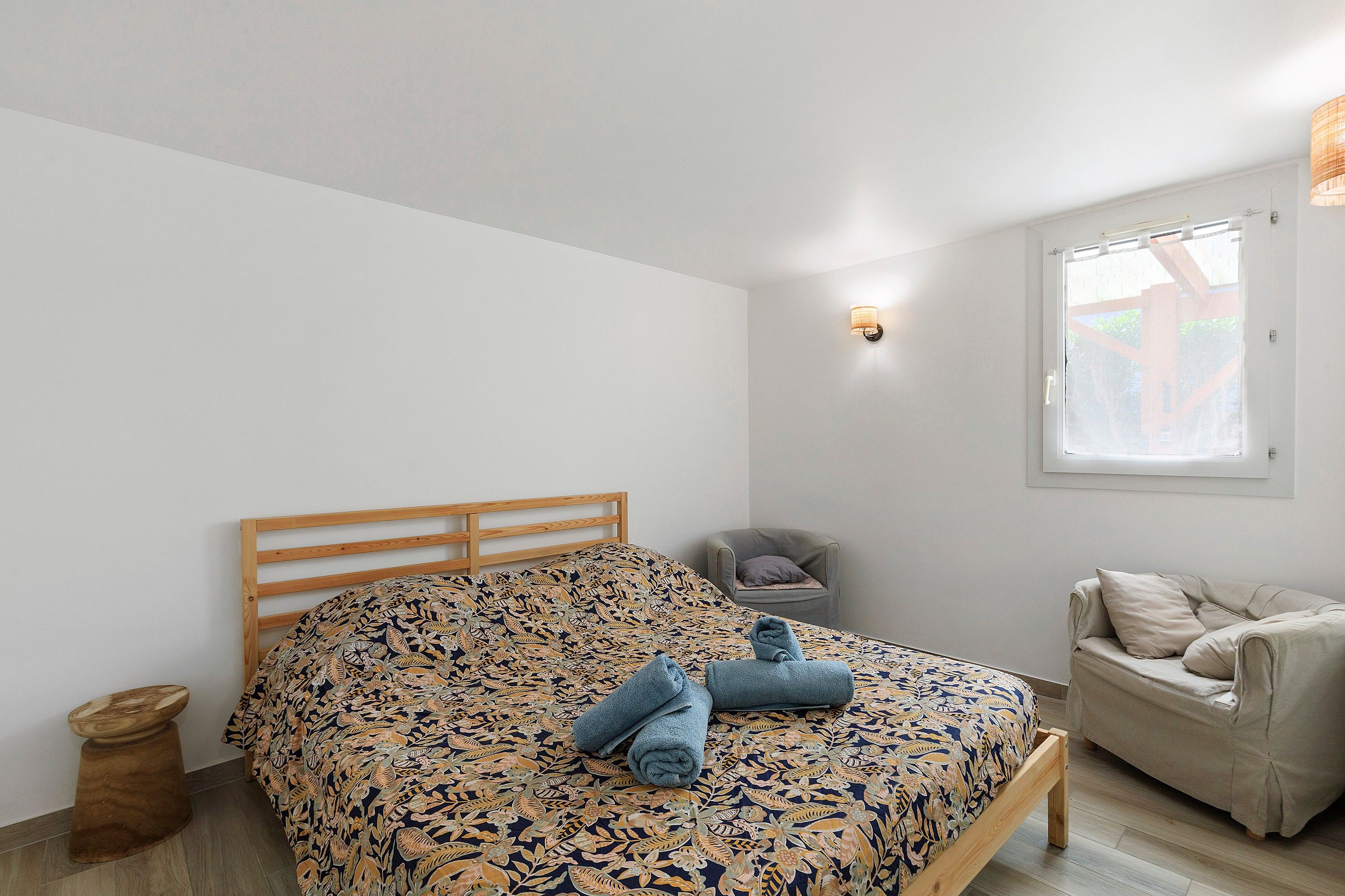 A bedroom with a wooden bed, a small bedside table, a chair, and a window with a view.