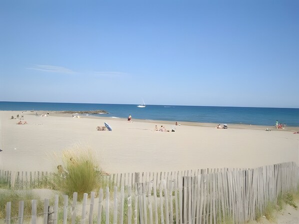 A beach with a wooden fence in the foreground, a sandy shore, and a sailboat in the distance.