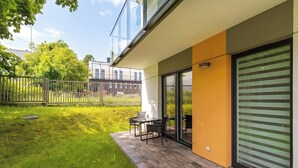 A modern outdoor patio with a table and chairs, a grassy area, and a building with large windows and a sliding door. - Cosy Apartment with Garden | Parking (Władysławowo)