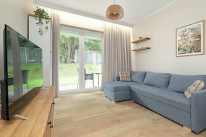 A modern living room with a flat-screen TV, a grey sectional sofa, wooden flooring, a large sliding glass door leading to a garden, and a woven pendant light. - Cosy Apartment with Garden | Parking (Władysławowo)