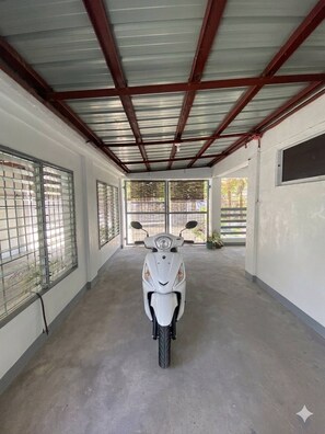 Ein überdachter Parkbereich mit einem weißen Roller in der Mitte, Fenstern mit Metallrahmen links und einem Betonboden.