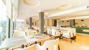 A modern dining area with white chairs and tables, a bar area with a green counter, and large windows offering a view.