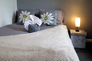 Ein ordentlich bezogenes Bett mit einer großen, strukturierten Decke, zwei Kissen mit Margeriten-Motiven, ein kleiner Nachttisch mit Lampe und ein мраморный Beistelltisch.
