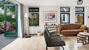 Ein modernes Wohnzimmer mit einer Couch, einer Bank, einem Tisch und einer Lampe. Es gibt Fenster mit Blick auf Bäume und ein Gebäude.