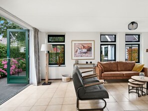 Ein modernes Wohnzimmer mit einer Couch, einer Bank, einem Tisch und einer Lampe. Es gibt Fenster mit Blick auf Bäume und ein Gebäude.