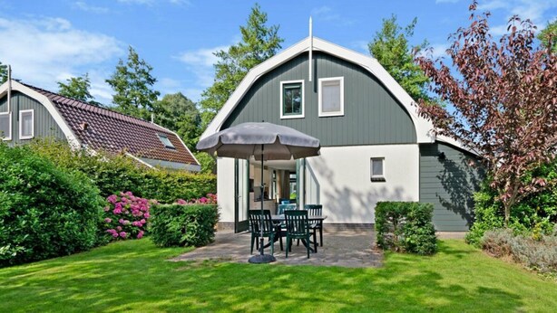 Ein Haus mit überdachter Terrasse, Rasen und einem Garten mit rosa Blüten.