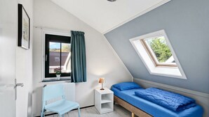 Ein Zimmer mit schrägen Wänden, einem Bett mit blauer Bettwäsche, einem weißen Stuhl, einem kleinen weißen Nachttisch, einem Fenster mit Vorhängen und einem Dachfenster.
