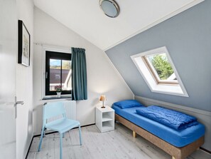 Ein Zimmer mit schrägen Wänden, einem Bett mit blauer Bettwäsche, einem weißen Stuhl, einem kleinen weißen Nachttisch, einem Fenster mit Vorhängen und einem Dachfenster.