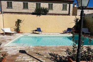 A rectangular swimming pool with two lounge chairs on each side, a stone building with green shutters in the background, and a street lamp in the foreground. - Apartment Camilla, Pesaro (Pesaro)
