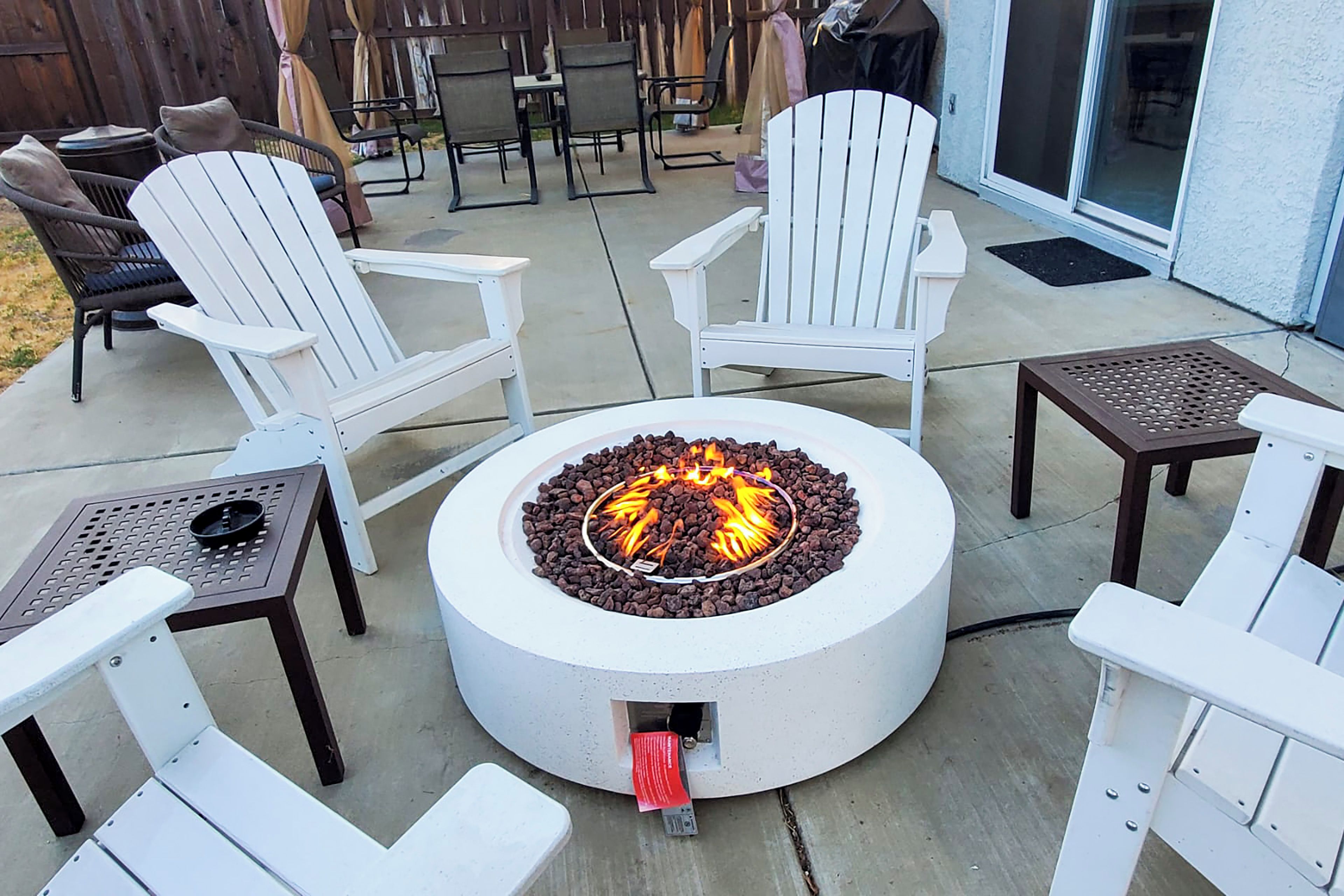 Un foyer circulaire, entouré de chaises Adirondack blanches et agrémenté d’une table avec un gril en métal.