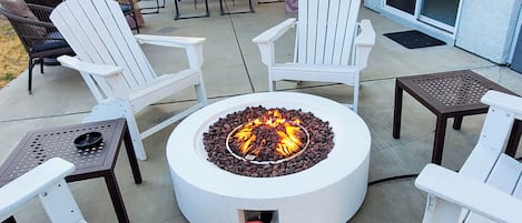 A circular fire pit surrounded by white Adirondack chairs and a table with a metal grill.