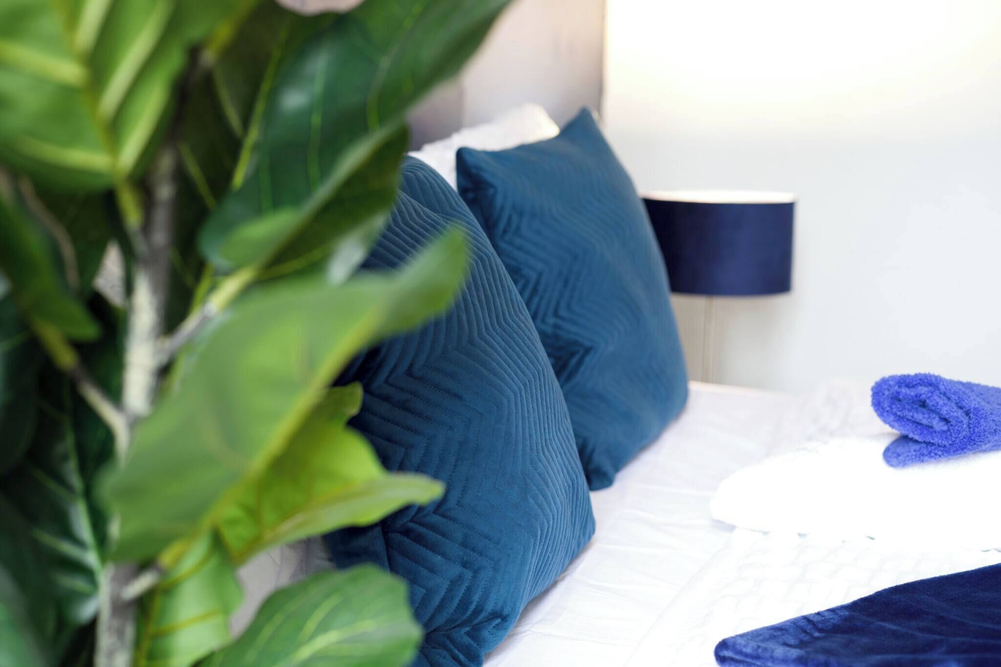 A bed with blue pillows, a folded blue towel, and a plant in the foreground.