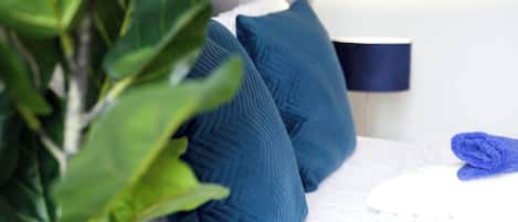 A bed with blue pillows, a folded blue towel, and a plant in the foreground.