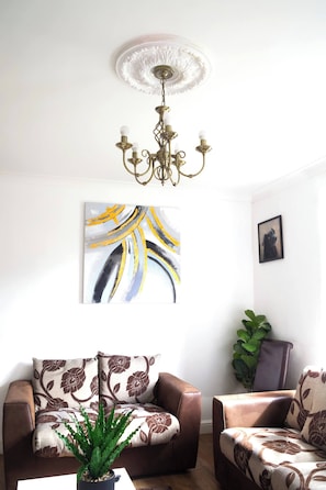 Una sala de estar con una araña, un sofá, un sillón, una planta en maceta y un cuadro en la pared.