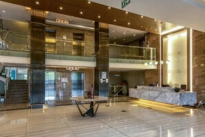 Een moderne hotellobby met een marmeren receptiebalie, een glazen lift en een trap.