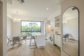 Une cuisine moderne équipée d’un lave-linge intégré, un coin repas avec une table en verre et une grande fenêtre donnant sur un paysage verdoyant.