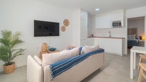 A modern living room with a sofa, a flat-screen TV, a kitchen area with cabinets and a microwave, and a dining table. - Cozy Apartment Rental in Guargacho, Spain - Ideal Family Stay (Guargacho)